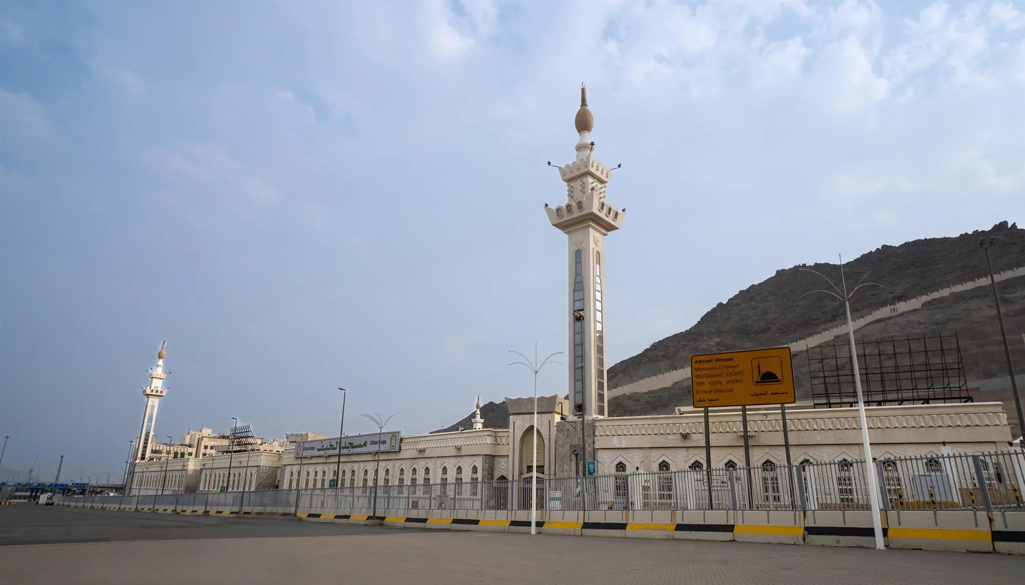 The exterior facade and a tall modern minaret of Masjid Al-Khayf in Makkah against the backdrop of the rugged mountains of Mina.