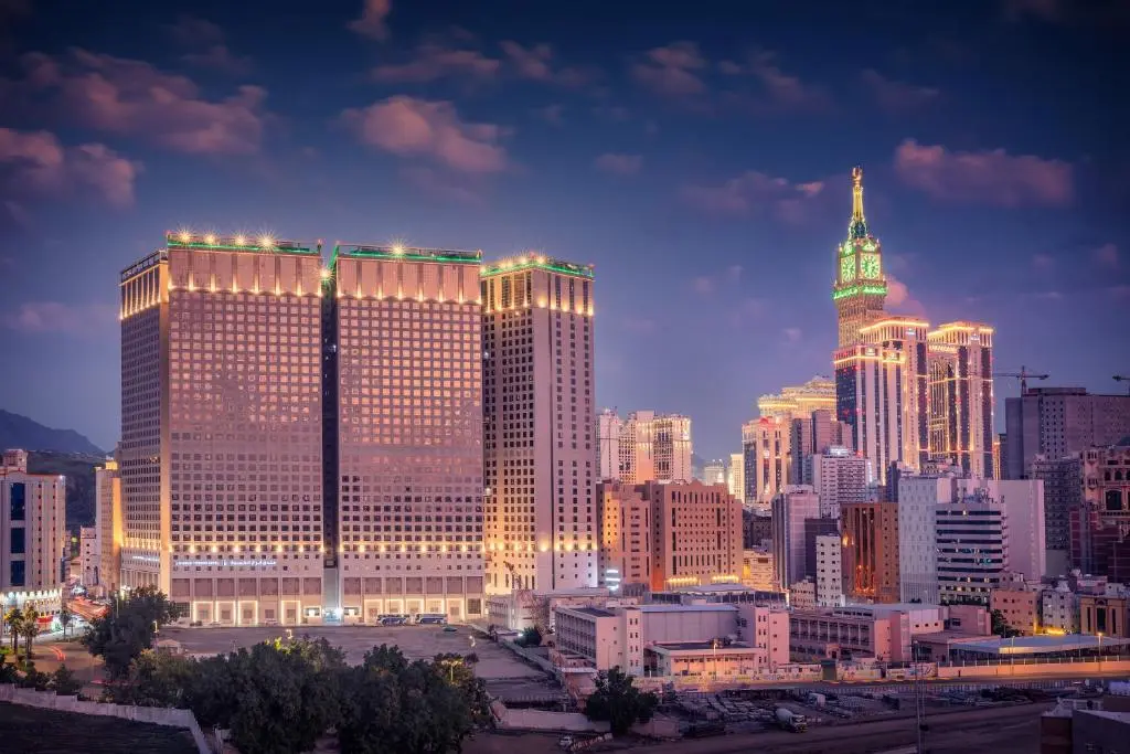 Night view of the Kiswa Towers in Makkah with the Clock Tower illuminated in the background.
