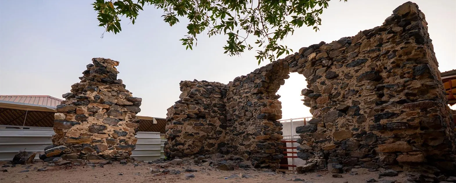 A wide view of the historical stone ruins and archway at the Hudaibiya site under a clear sky.